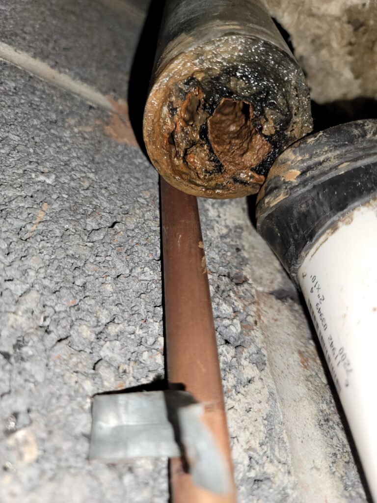 Cut end of a corroded drain pipe packed with rust scale, next to a copper line and a white PVC pipe